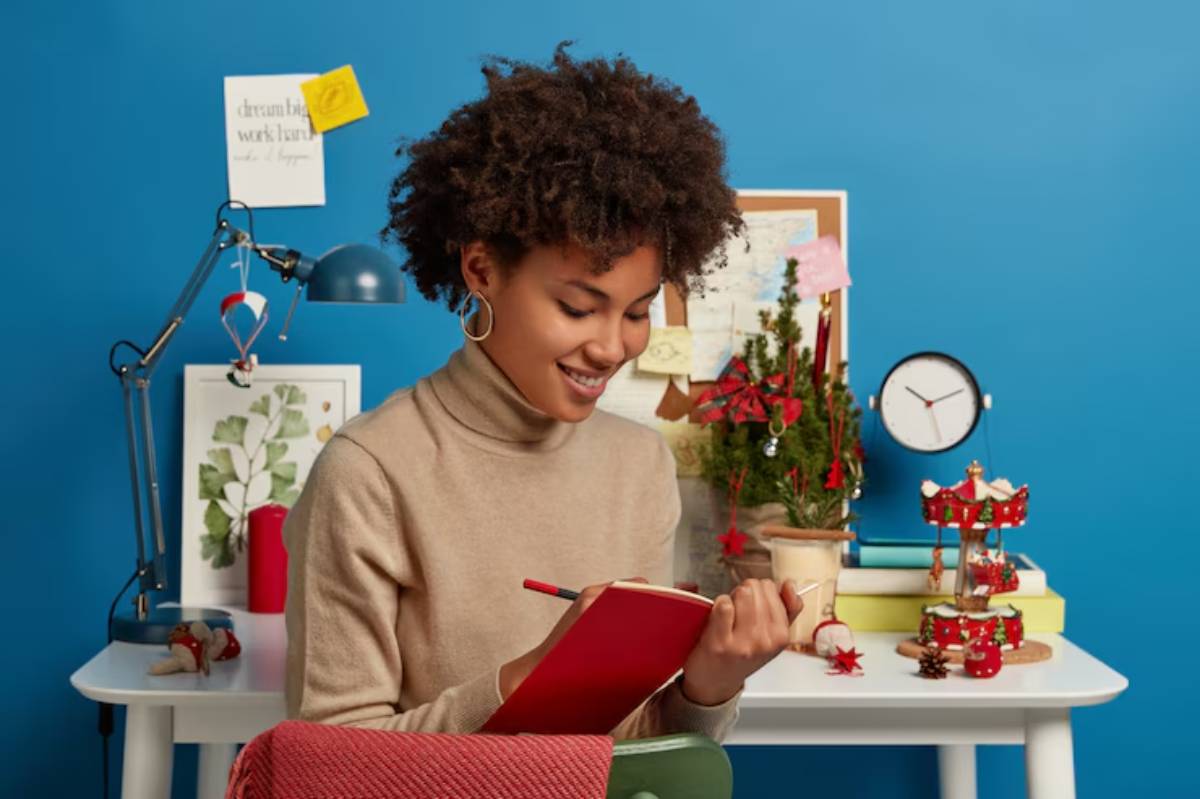 A person holding a red notebook sits at a desk adorned with holiday decorations, plants, and colorful wall art against a blue background.