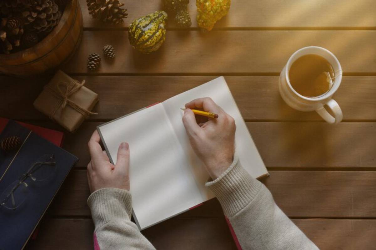 A person writes in a blank notebook with a yellow pencil, surrounded by pinecones, gourds, a cup of tea, and gifts on a wooden table.