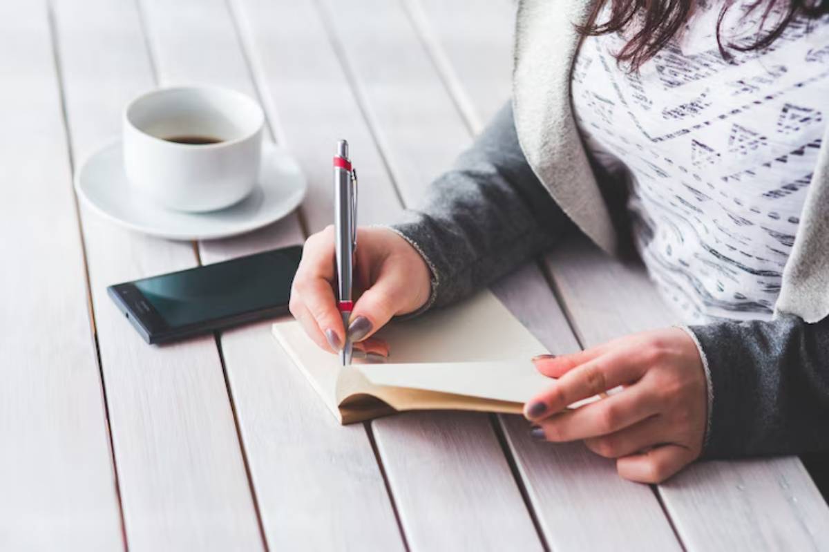 A person writes in a blank notebook while seated at a table with a cup of coffee and a smartphone nearby.