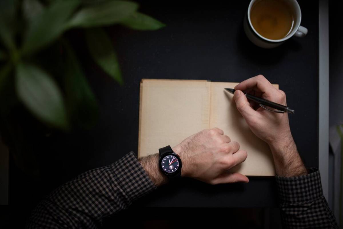 A hand writes in a blank notebook with a pen, next to a cup of tea and surrounded by a green plant.