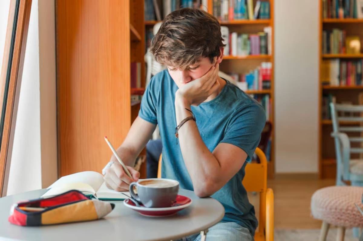 A young person sits at a table in a cozy library cafe, writing in a notebook with a cup of coffee nearby. Shelves filled with books are visible.