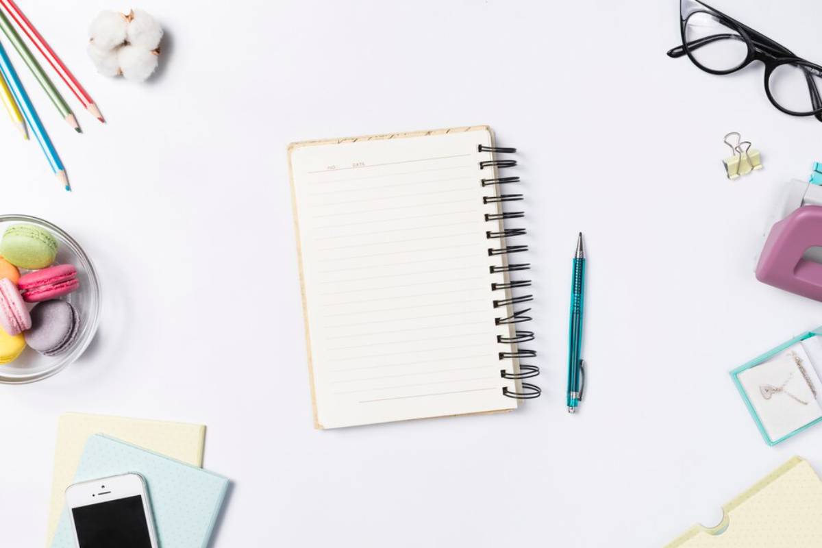 An empty notebook surrounded by colorful pencils, macarons, glasses, a phone, stationery, and a jewelry box on a white background.