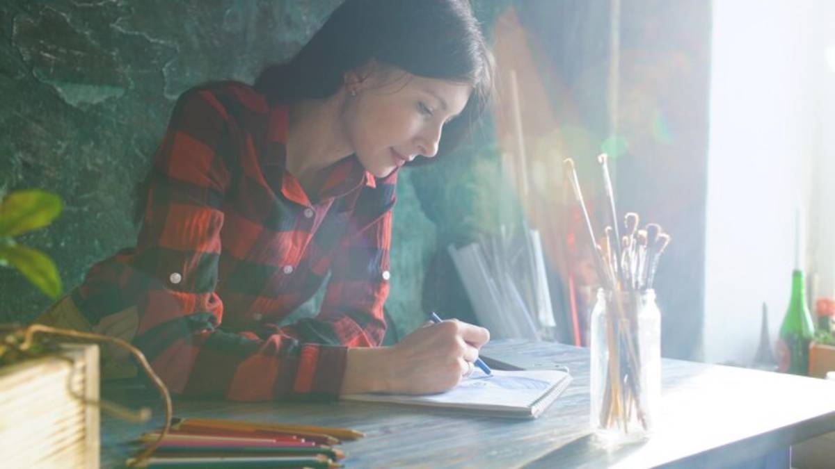 A person in a red plaid shirt sketches in a notebook, surrounded by art supplies, on a sunlit table with a green backdrop.