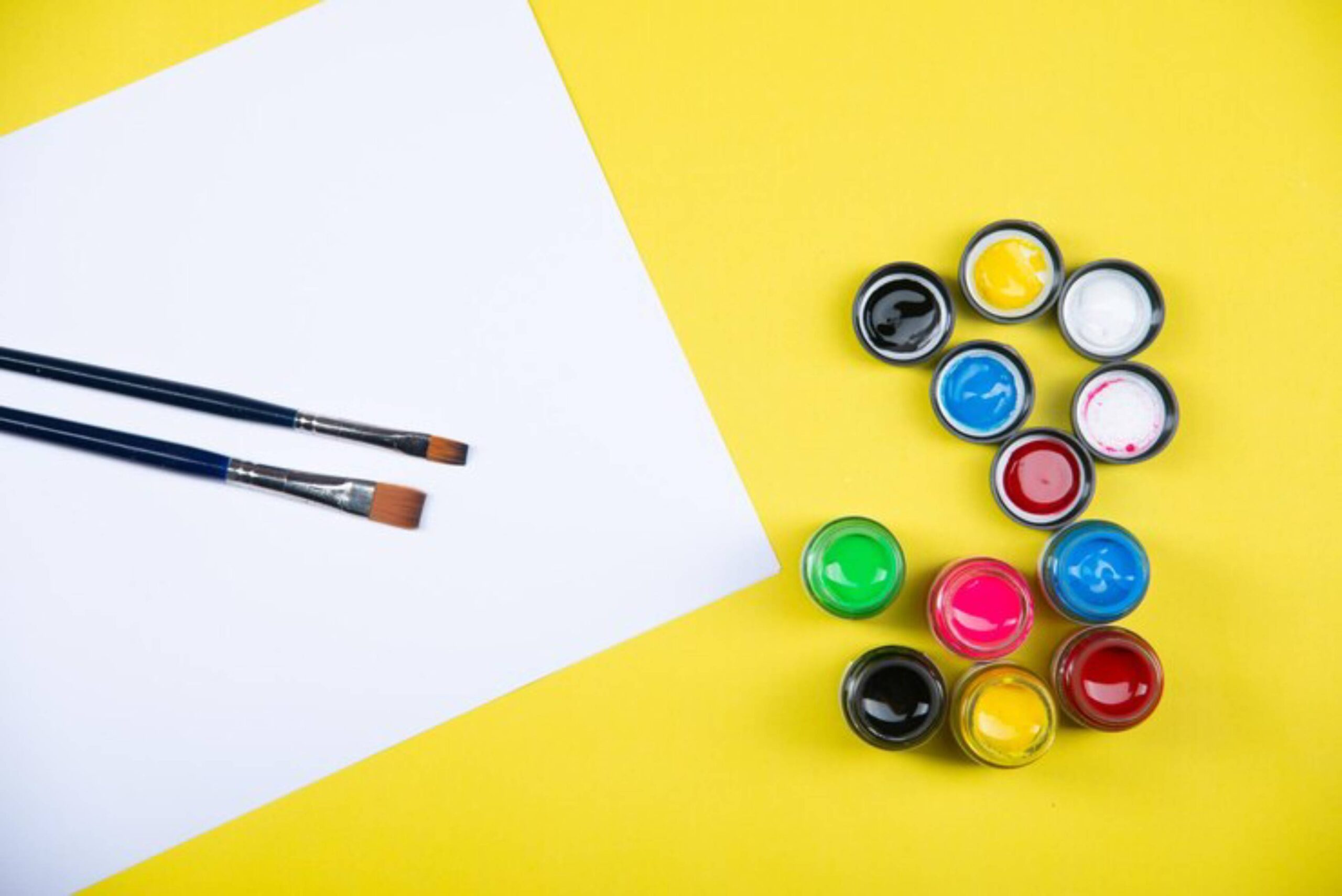 Two paintbrushes and a blank sheet of paper beside colorful paint pots on a bright yellow background.