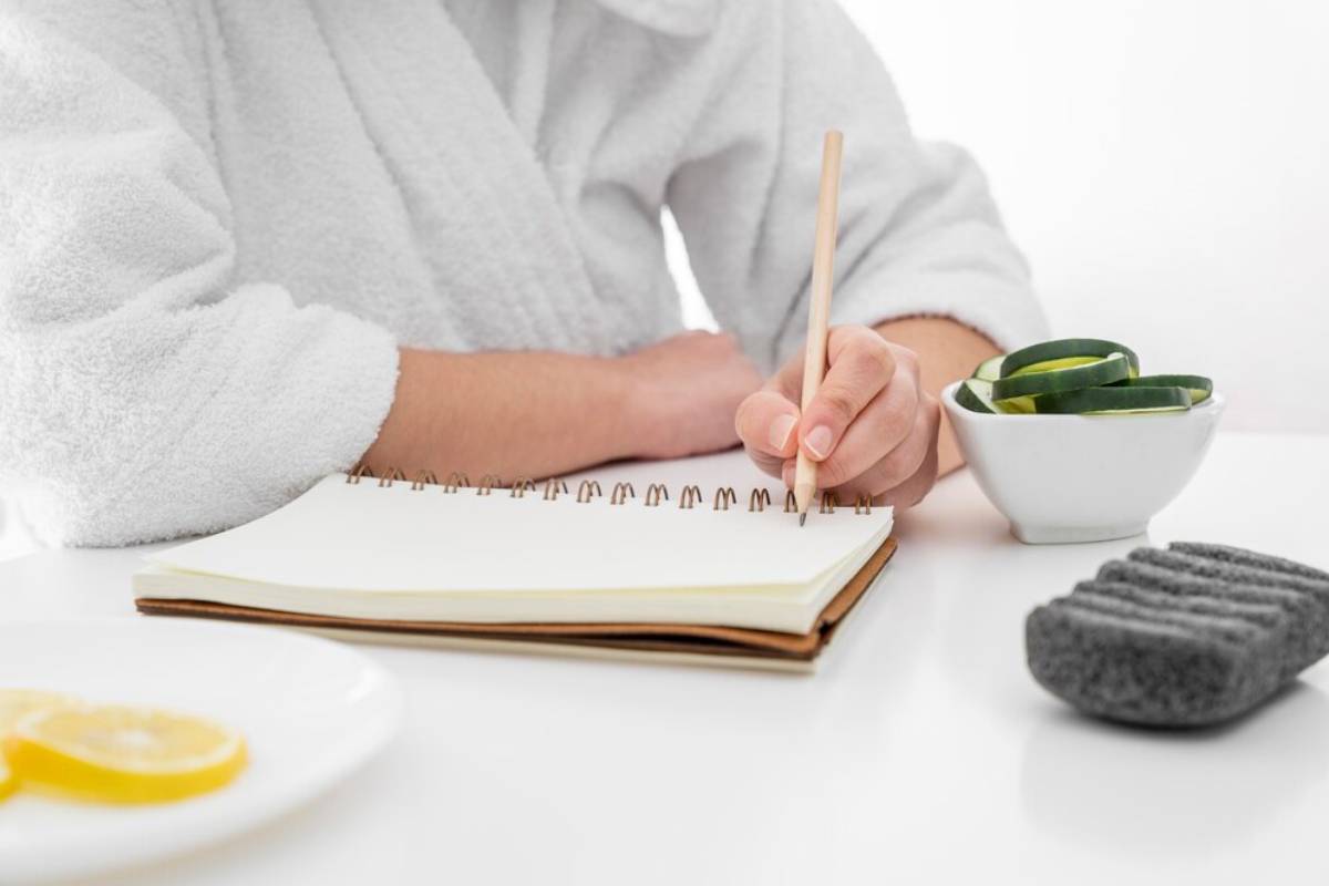 A person in a white robe writes in a notebook, surrounded by slices of lemon, cucumber, and skincare stones on a white table.