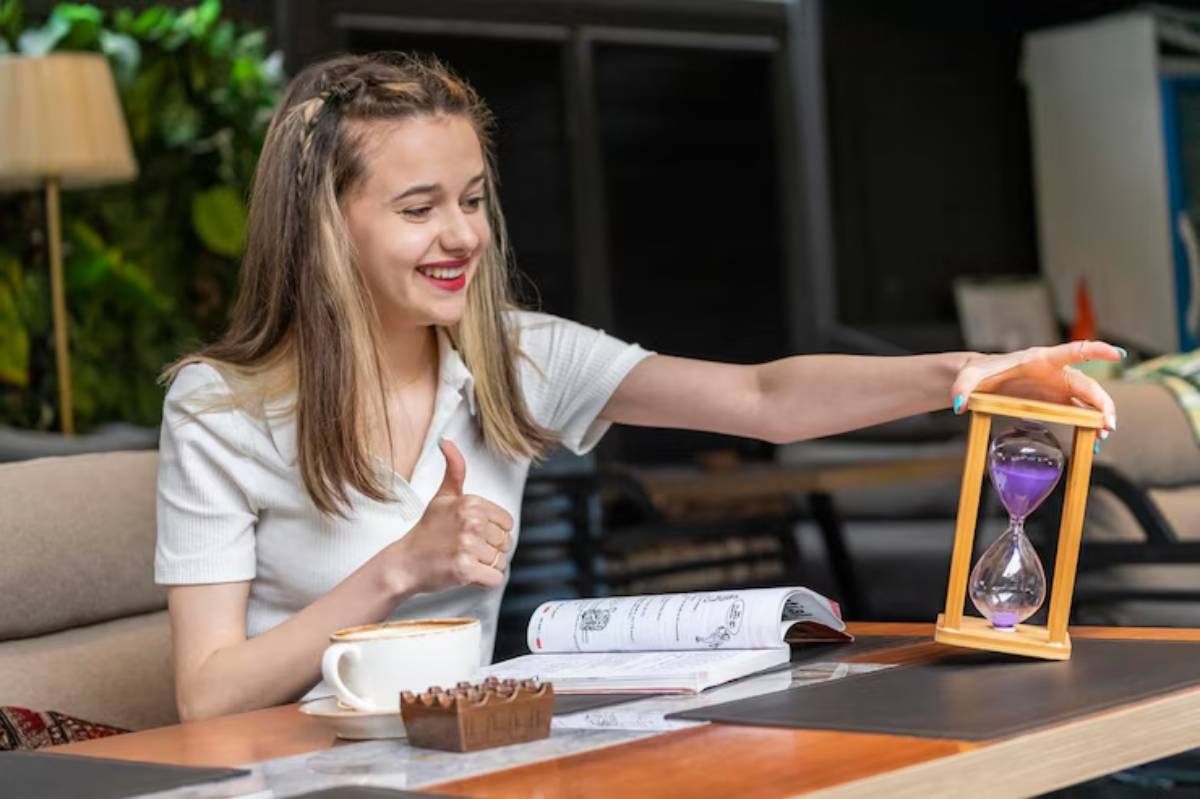 A young woman sitting at a table, giving a thumbs up, holding an hourglass. Coffee cup and an open book are on the table.