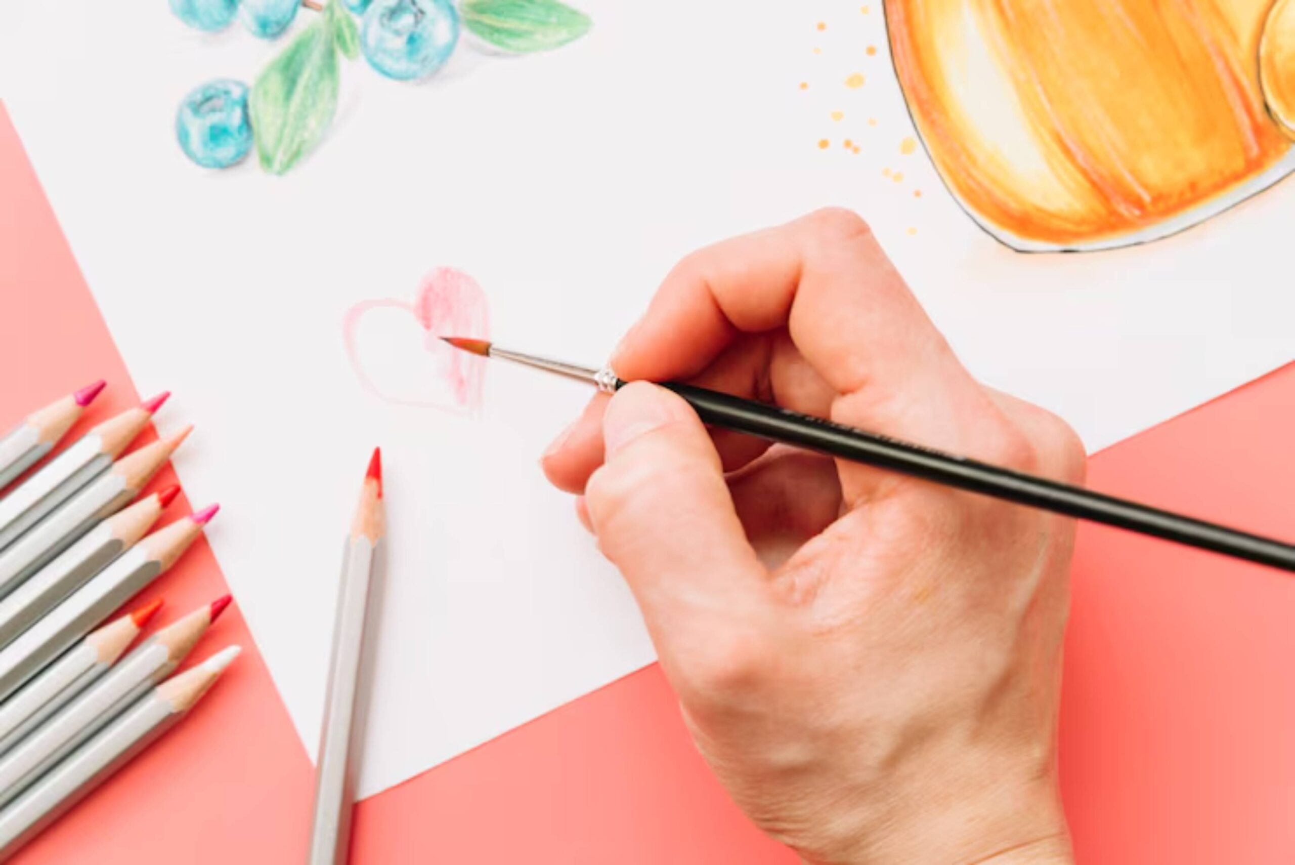 A hand holds a paintbrush, adding pink strokes to a heart shape on paper, surrounded by colored pencils and artistic sketches.