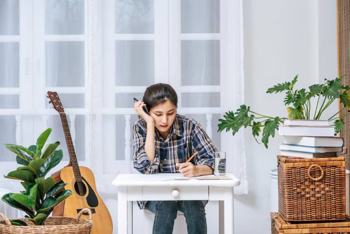 A person sits at a white table, taking notes with a pencil, surrounded by plants, a guitar, and stacked books in a bright room.
