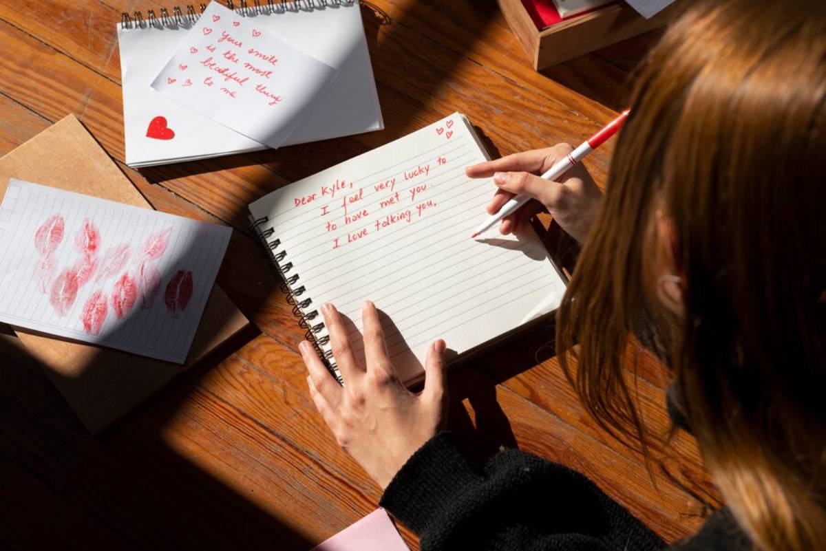 A person writing a love letter in a notebook, surrounded by lips prints, notes, and a warm wooden table setting.