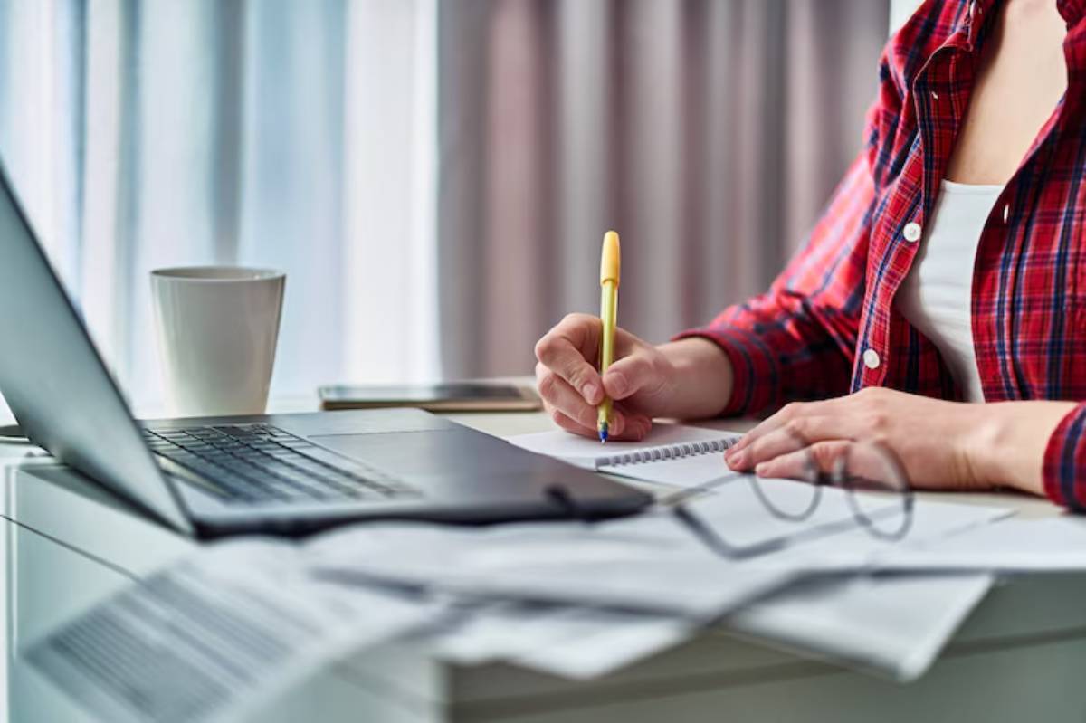 A person in a plaid shirt writes notes in a notebook while sitting at a desk with a laptop and a coffee mug nearby.