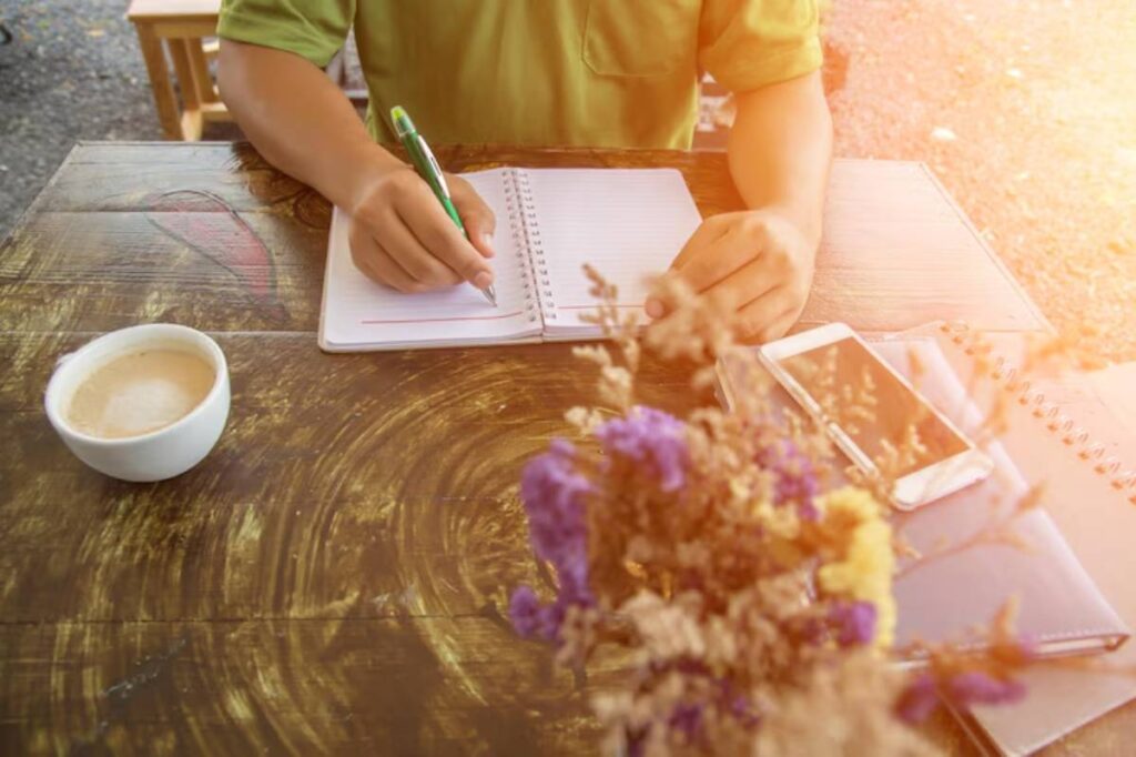 A person writes in a notebook with a green pen, accompanied by a cup of coffee and a smartphone on a wooden table.