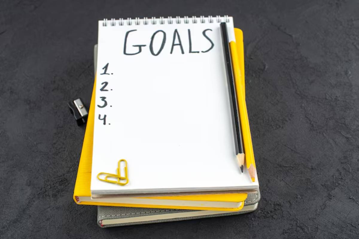 A blank notepad titled GOALS with numbered lines, a black pencil, a yellow pencil, and paper clips on a textured black background.