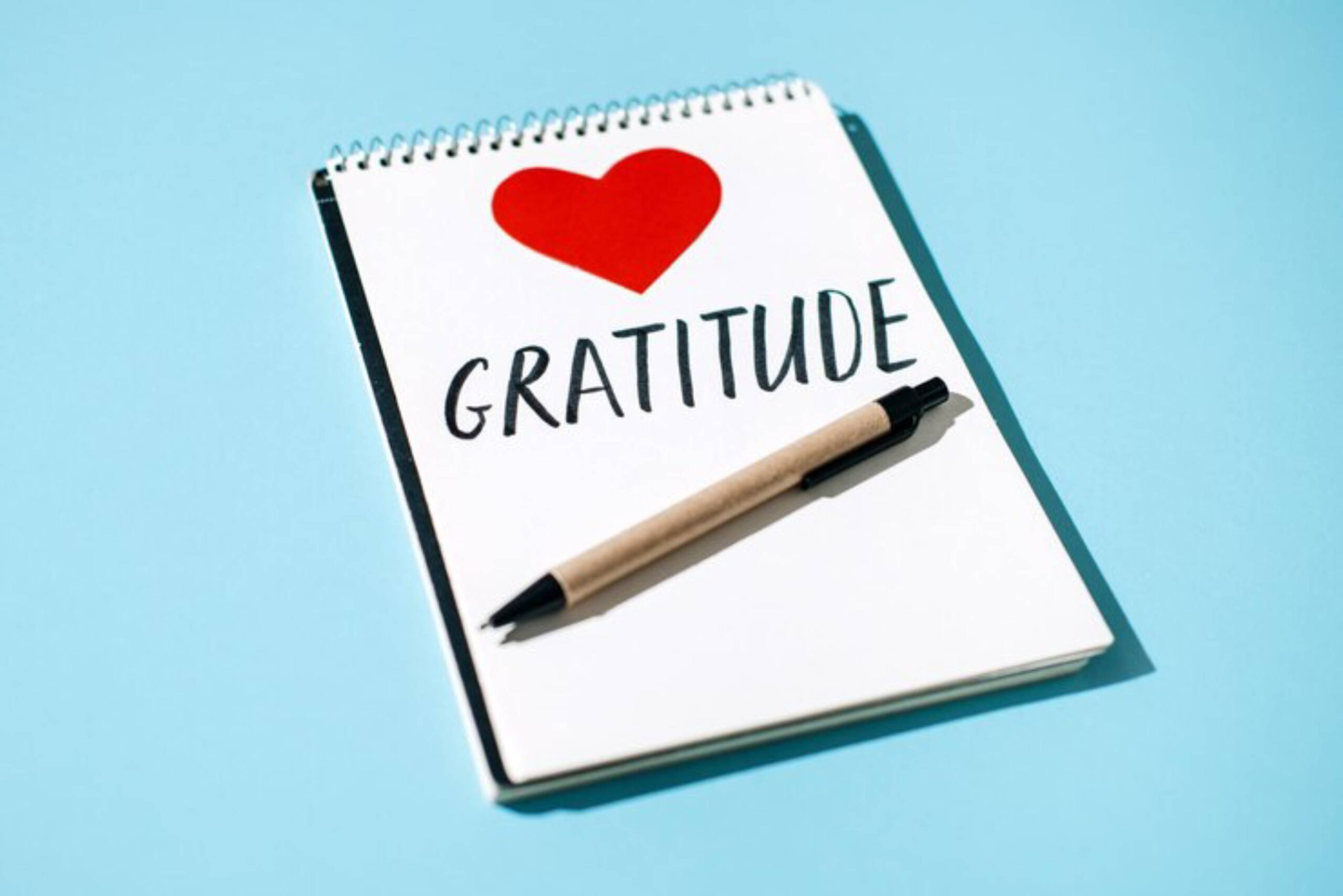 A spiral-bound notebook with GRATITUDE written in bold and a red heart above it, accompanied by a black pen on a light blue background.