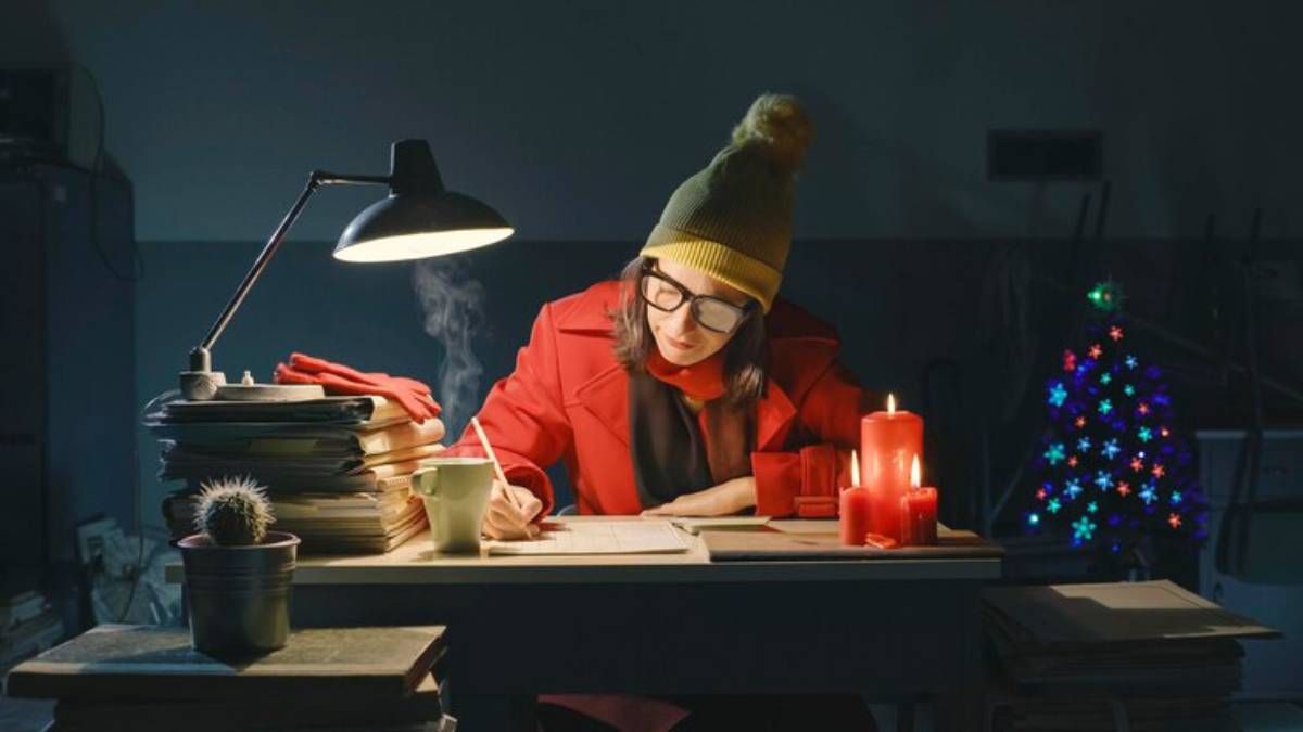 A person in a red coat and yellow beanie writes at a desk surrounded by stacks of papers, candles, and a small decorated Christmas tree.