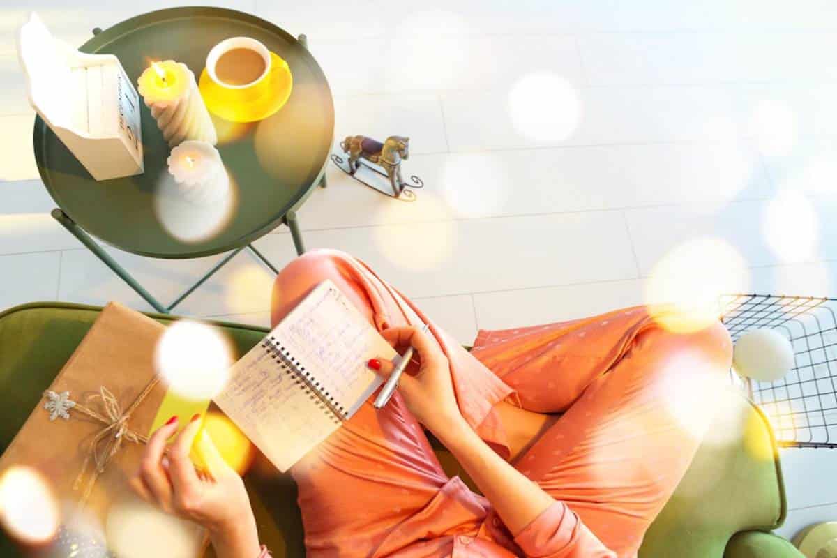 A person in pink pajamas sits on a green couch, writing in a notebook beside a table with candles and a coffee cup. Warm, cozy atmosphere.