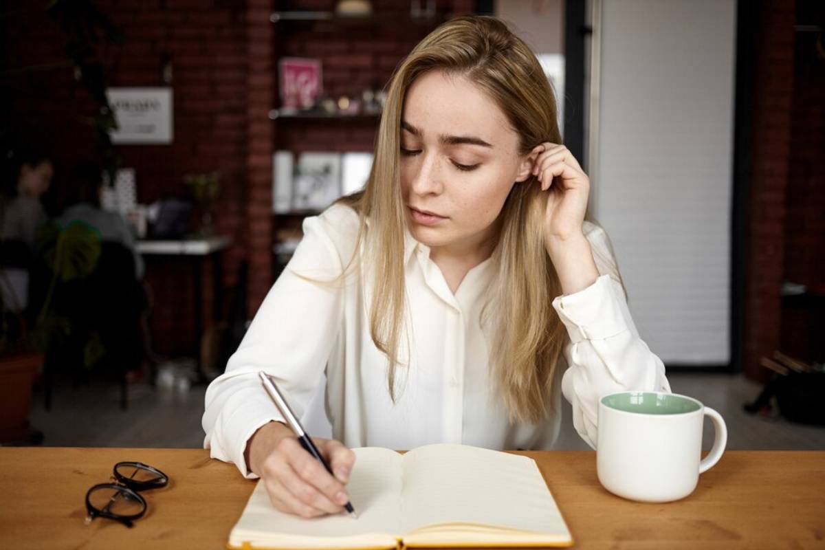 A person in a white shirt writes in a notebook at a wooden table, with a mug and glasses nearby, in a cozy workspace.