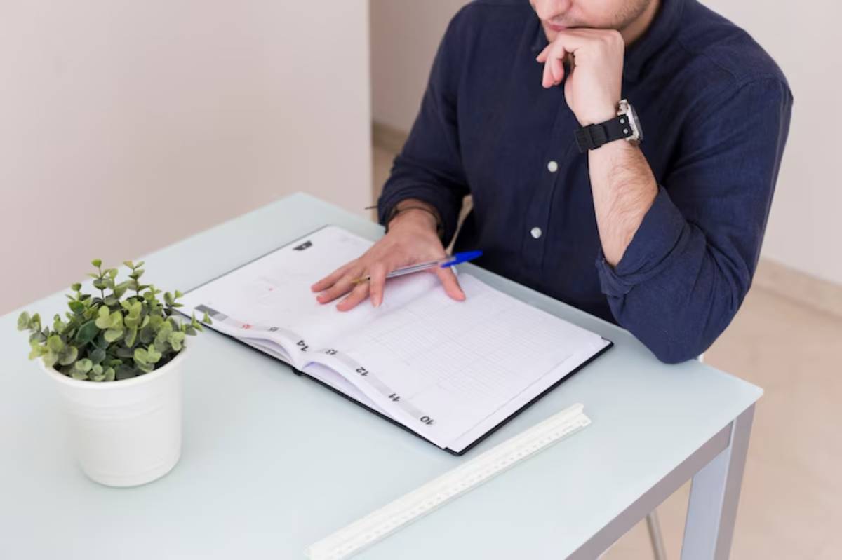 A man in a dark shirt sits at a desk, contemplating while writing in a planner with a blue pen, a small plant nearby.