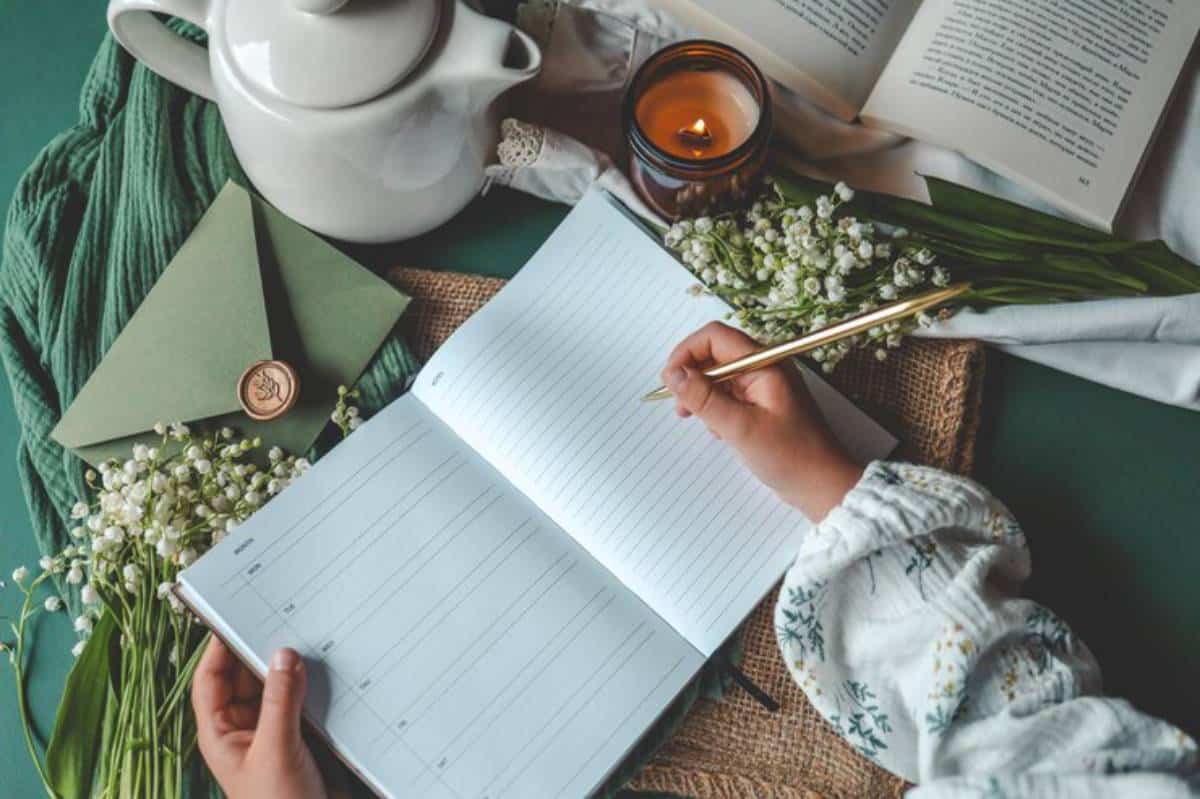 A hand holds a gold pen over a blank notebook, surrounded by flowers, a green envelope, a teapot, and a lit candle.