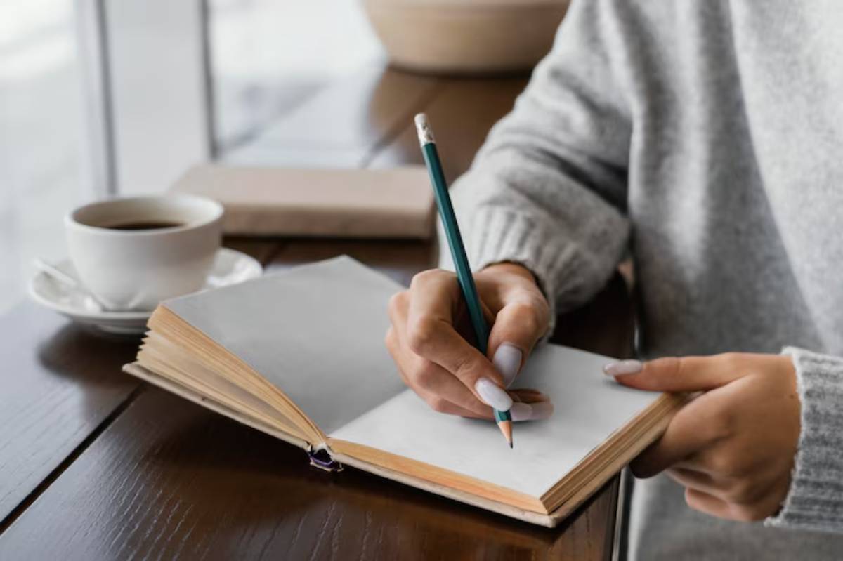 A person in a cozy sweater writes in a blank notebook, with a cup of coffee nearby on a wooden table.