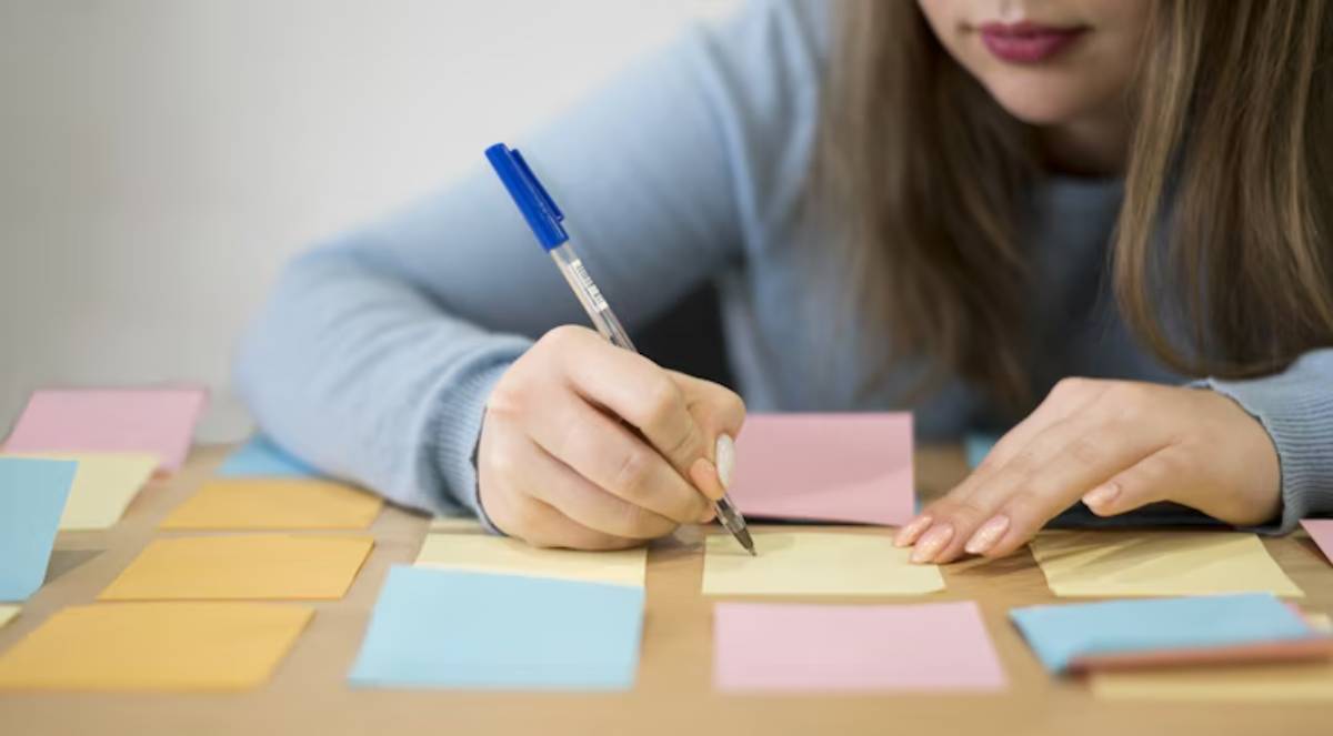 A woman in a light blue sweater writes on a yellow sticky note surrounded by colorful notes on a wooden table.