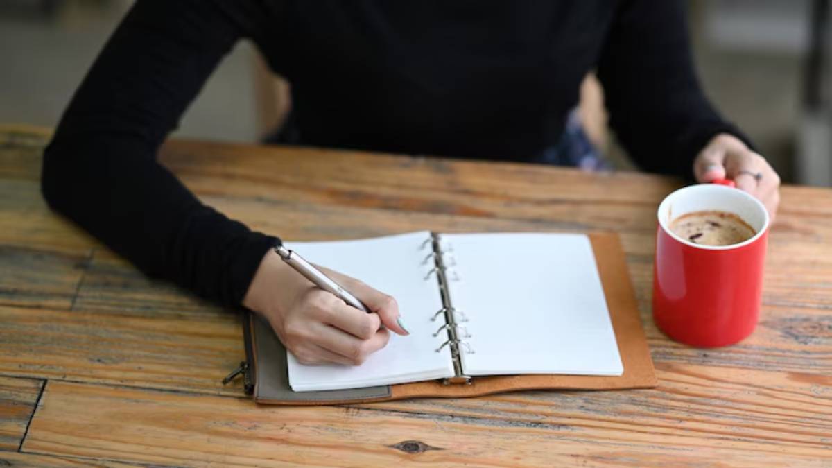 A person's hand writes in a notebook while holding a red coffee mug, set on a rustic wooden table.