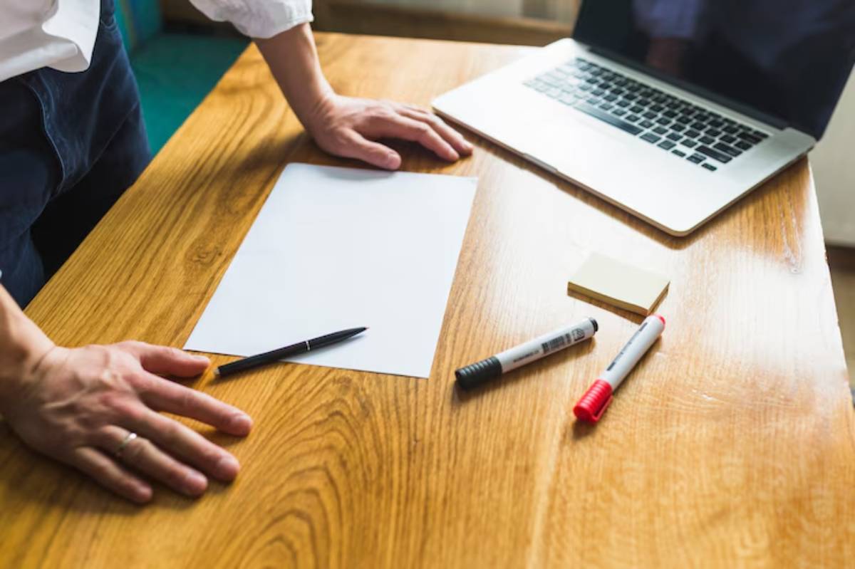 A person's hands rest on a wooden desk with a laptop, blank paper, pen, sticky notes, and markers scattered around.