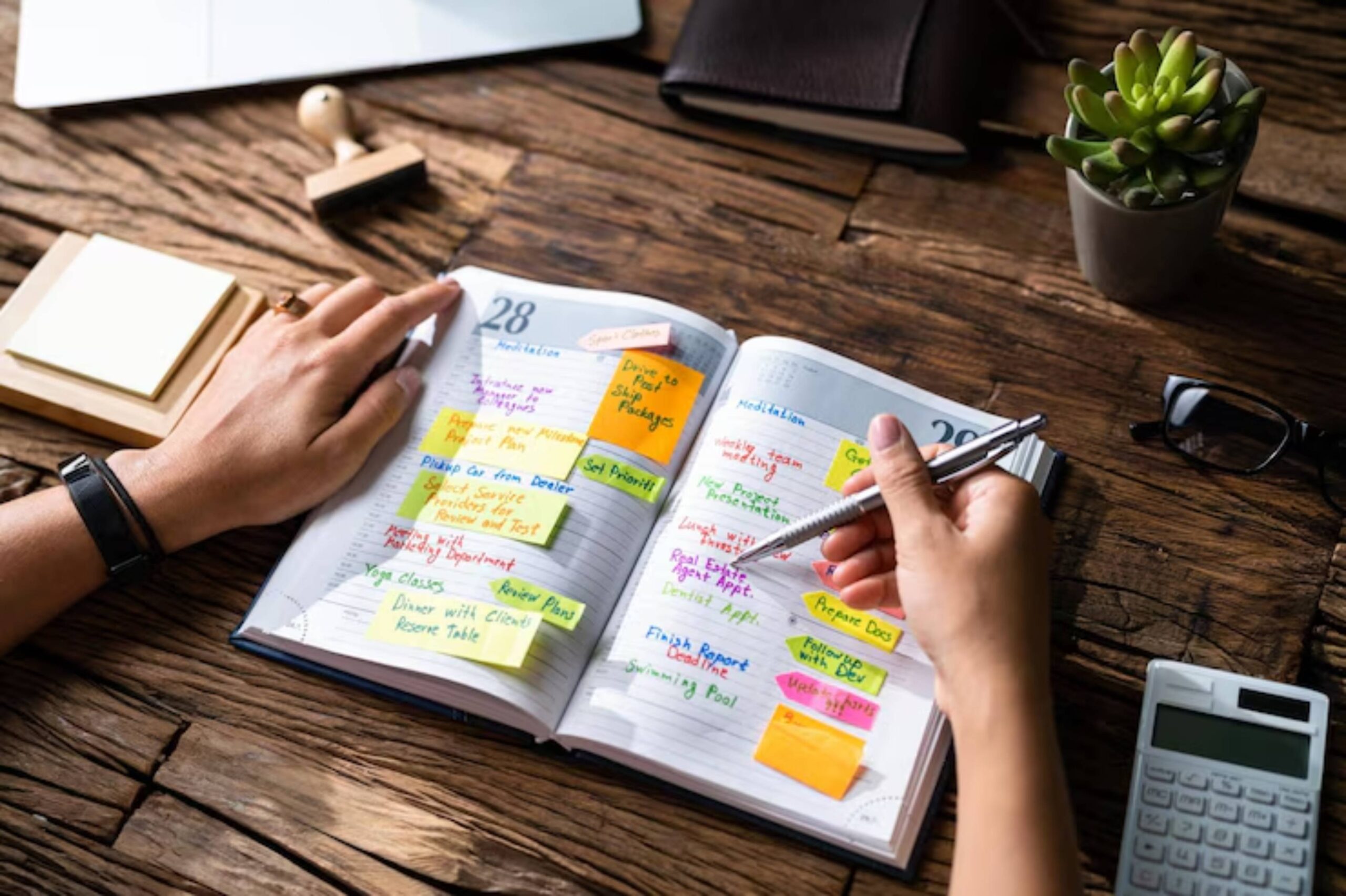 A hand holds a pen over a planner filled with colorful sticky notes and tasks, alongside a laptop, calculator, and a plant.