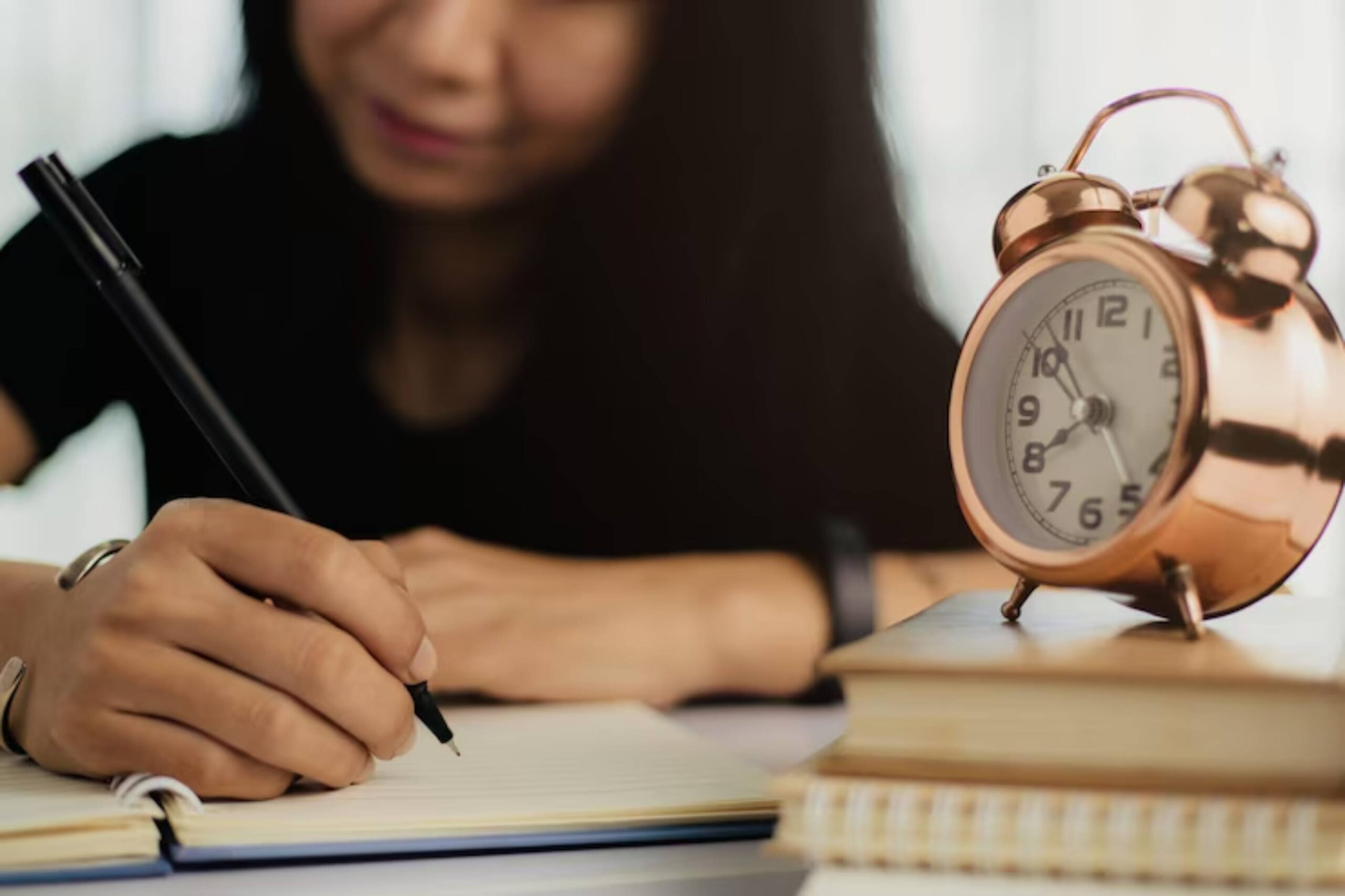 A person writes in a notebook with a black pen, while a vintage alarm clock sits atop a stack of books in the foreground.