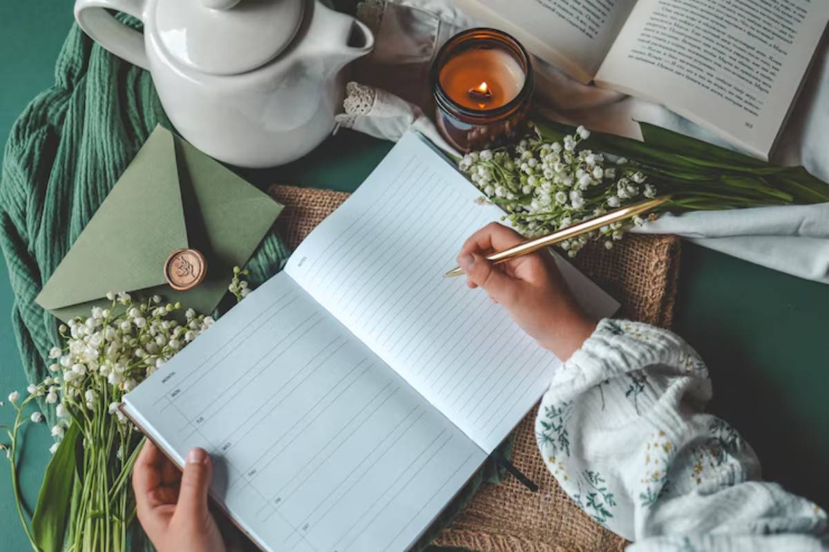 A hand with a gold pen writes in a blank notebook, surrounded by flowers, a teapot, a candle, and a green envelope on a textured surface.