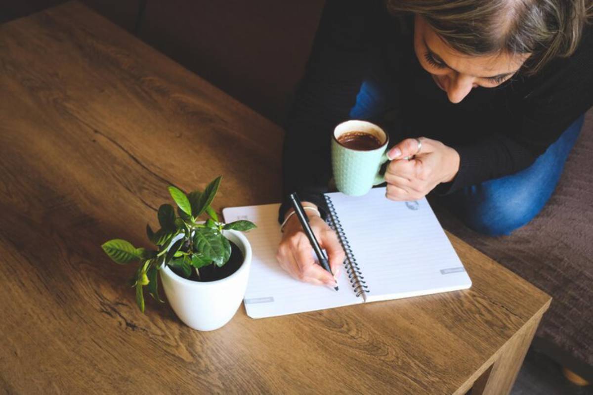 A person's hand holding a pen writes in a notebook on a wooden table, alongside a small potted plant and a cup of coffee.