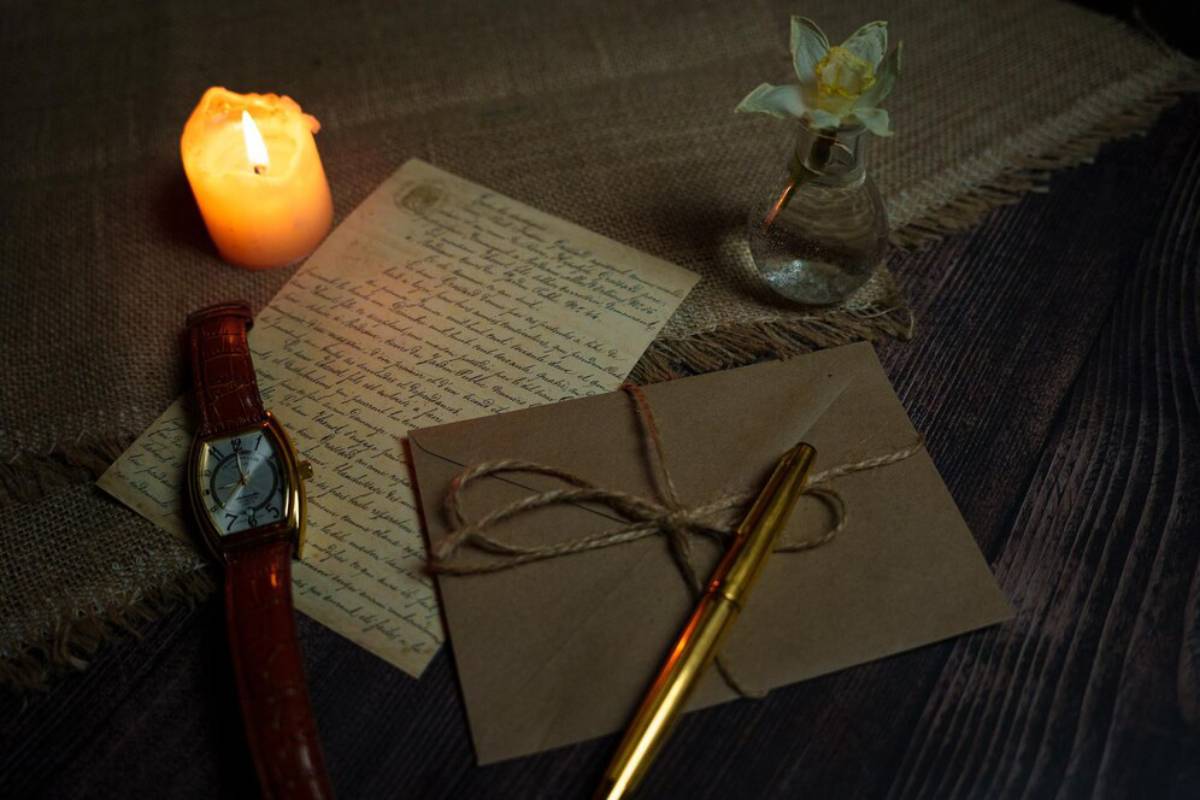 A candle illuminates a vintage letter, a watch, a tied envelope, and a pen, evoking a nostalgic, warm ambiance on a rustic table.