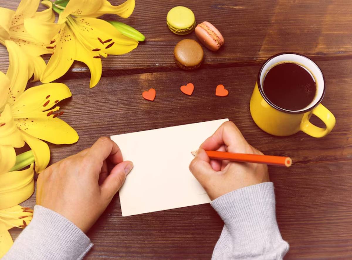 A hand holding a colored pencil writes on a blank card surrounded by yellow lilies, macarons, and a cup of coffee on a wooden table.