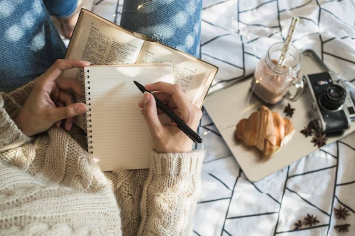 A cozy scene featuring a person writing in a notebook, surrounded by a book, a cup of chocolate drink, a croissant, and a vintage camera.