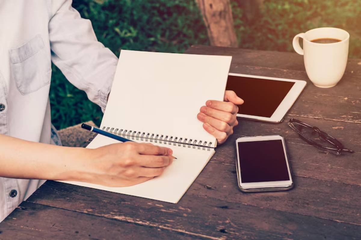 A hand writes in a blank notebook, surrounded by a smartphone, tablet, glasses, and a coffee cup on a rustic table.