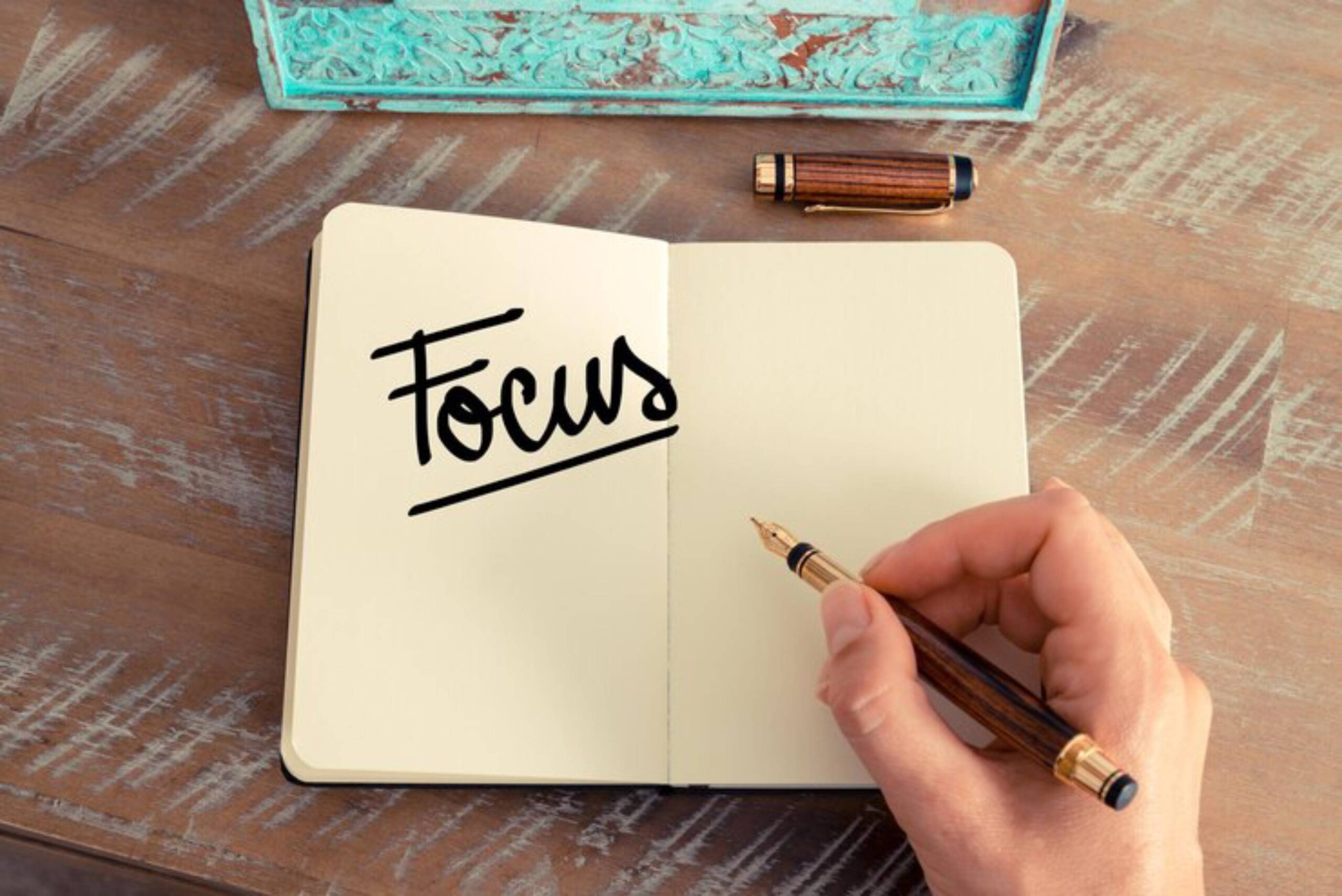 A hand holds a fountain pen poised over an open notebook, which prominently displays the word 