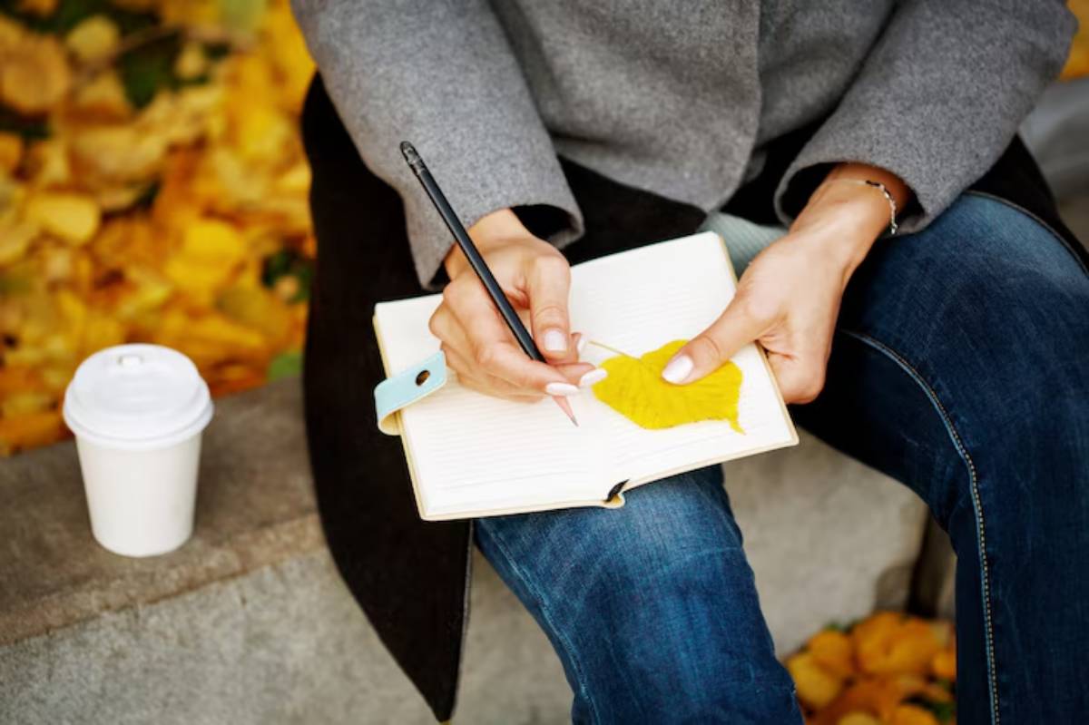 A person in a gray coat sketches a yellow leaf in a notebook while sipping coffee, surrounded by autumn leaves.