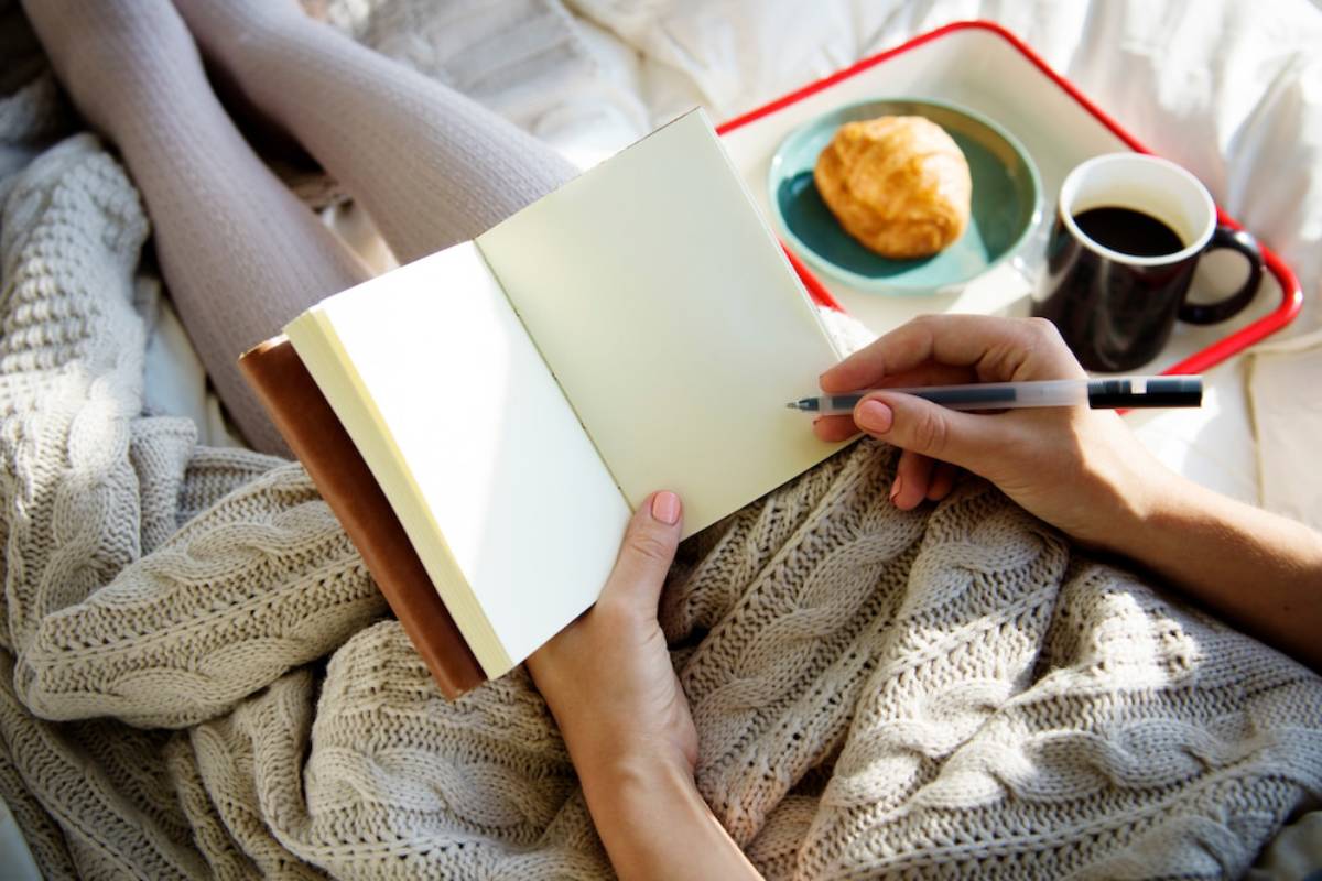 A person writes in a blank notebook while seated on a cozy blanket, with a cup of coffee and a croissant on a tray nearby.