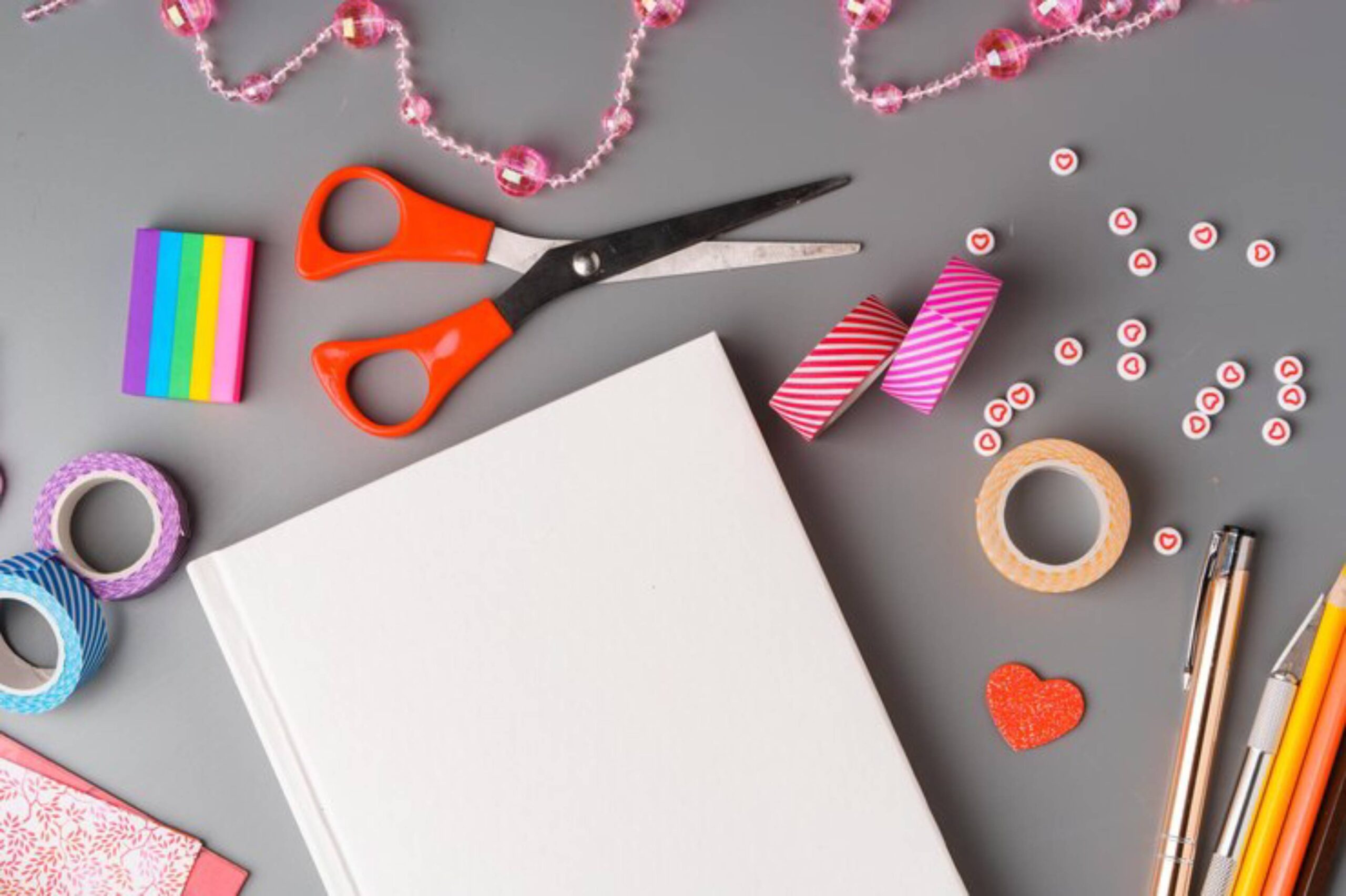 An array of crafting supplies including scissors, colorful tape, an empty notebook, and decorative beads arranged on a gray background.