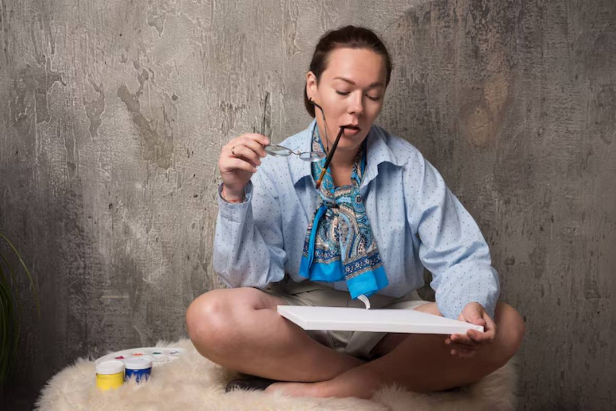A seated person in a blue blouse with a patterned scarf holds a sketchbook, surrounded by paint containers against a textured wall.
