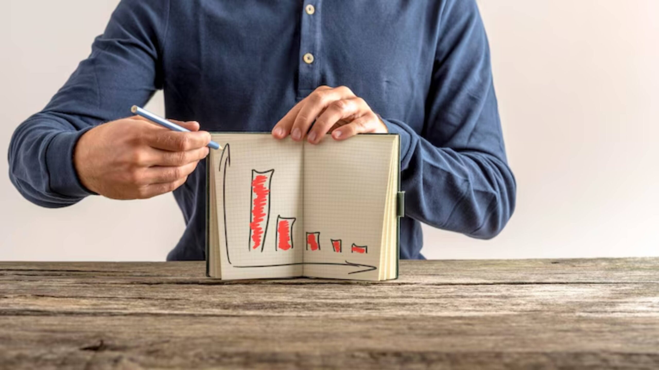A person in a blue shirt demonstrates a hand-drawn bar graph in a notebook, indicating trends with a pencil on a wooden table.