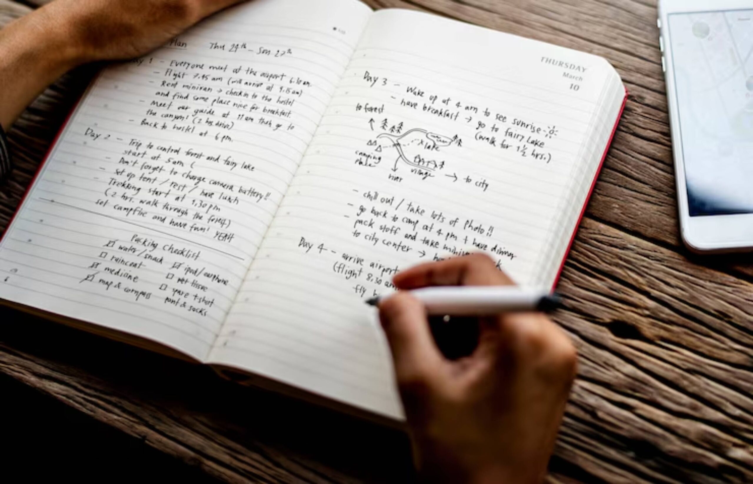A hand writing in a notebook with travel plans, to-do lists, and a packing checklist, alongside a smartphone on a wooden table.