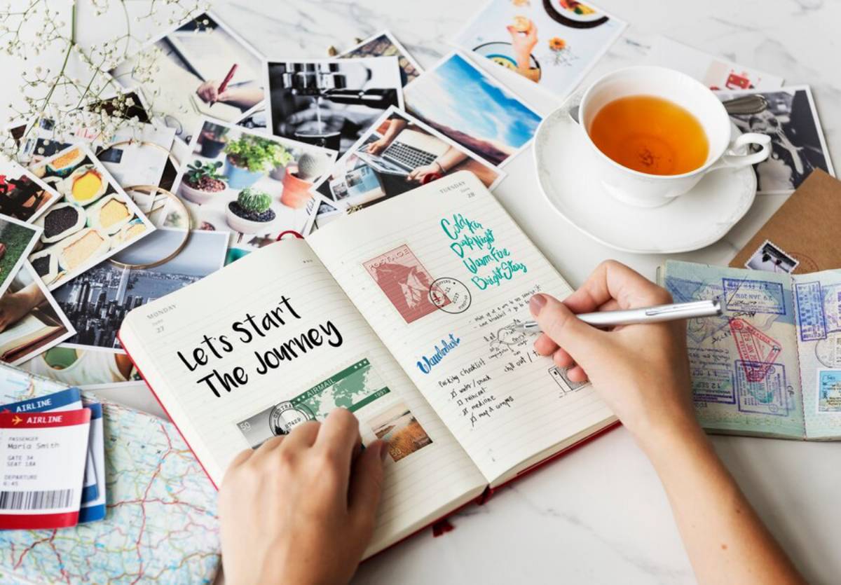 A hand writes in a travel journal titled Let's Start The Journey, surrounded by photos, a cup of tea, and travel documents.