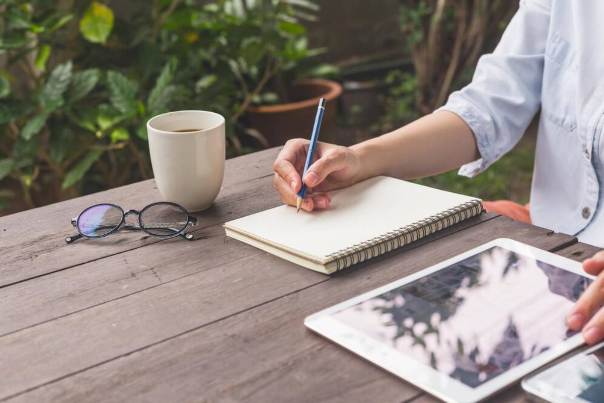 A person writes in a notebook with a pencil, a coffee cup, and glasses nearby, surrounded by green plants on a wooden table.