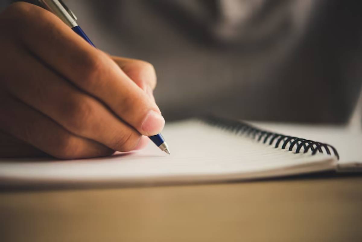 A close-up of a hand holding a blue pen, writing in a spiral-bound notebook with blank pages.