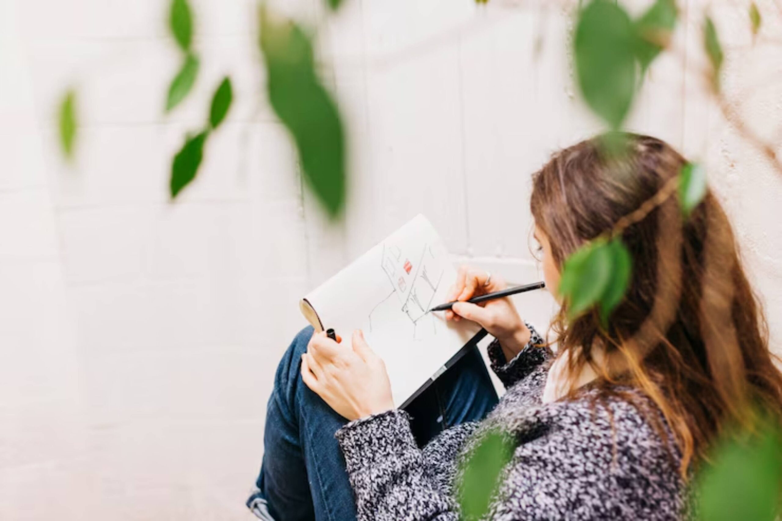 A woman sketches in a notebook while sitting against a wall, partially obscured by green leaves.