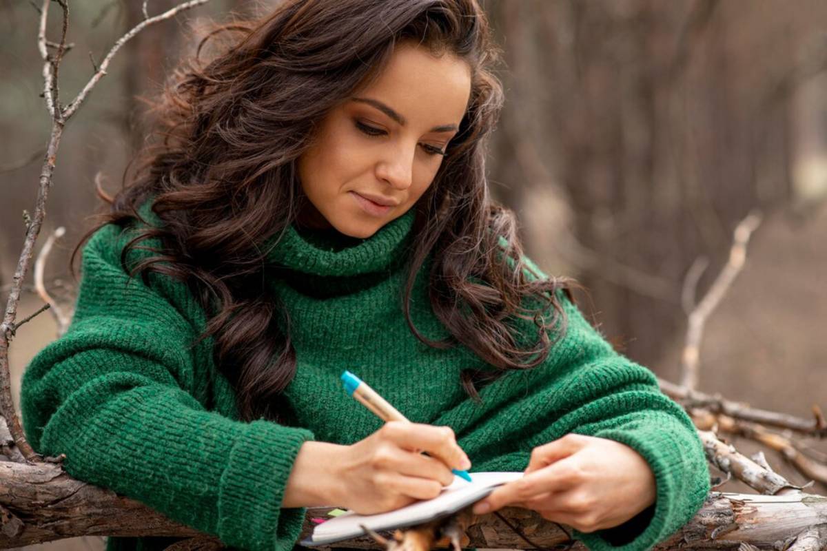 A person in a cozy green sweater writes in a notebook, surrounded by natural, blurred trees and branches.