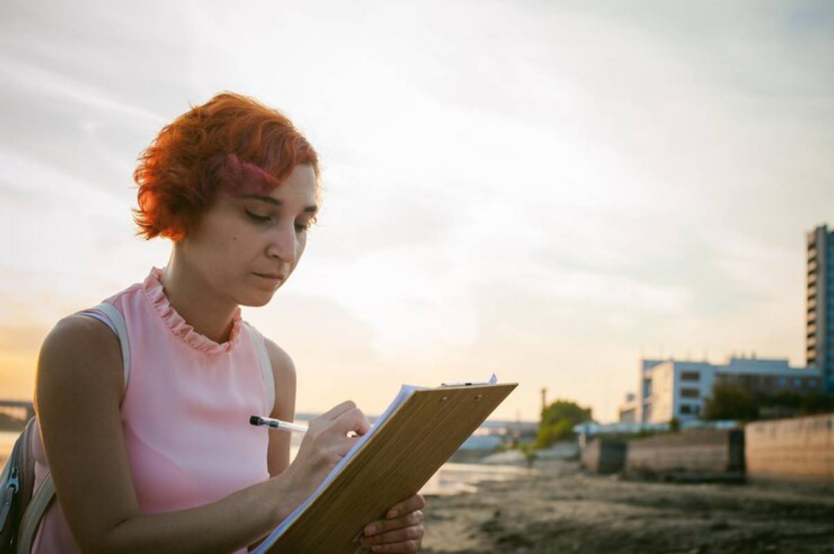 A woman with bright red hair writes on a clipboard by the waterfront during sunset, surrounded by urban buildings and greenery.