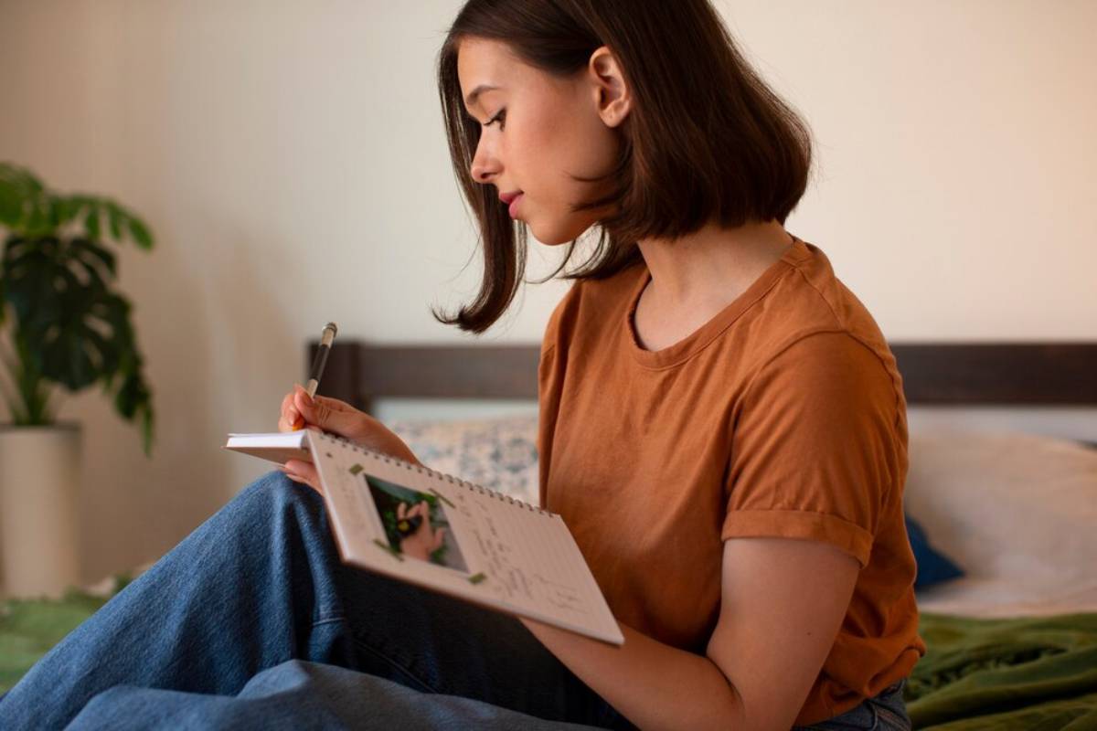 A person sits on a bed, wearing an orange shirt, writing in a notebook. A green plant is visible in the background.