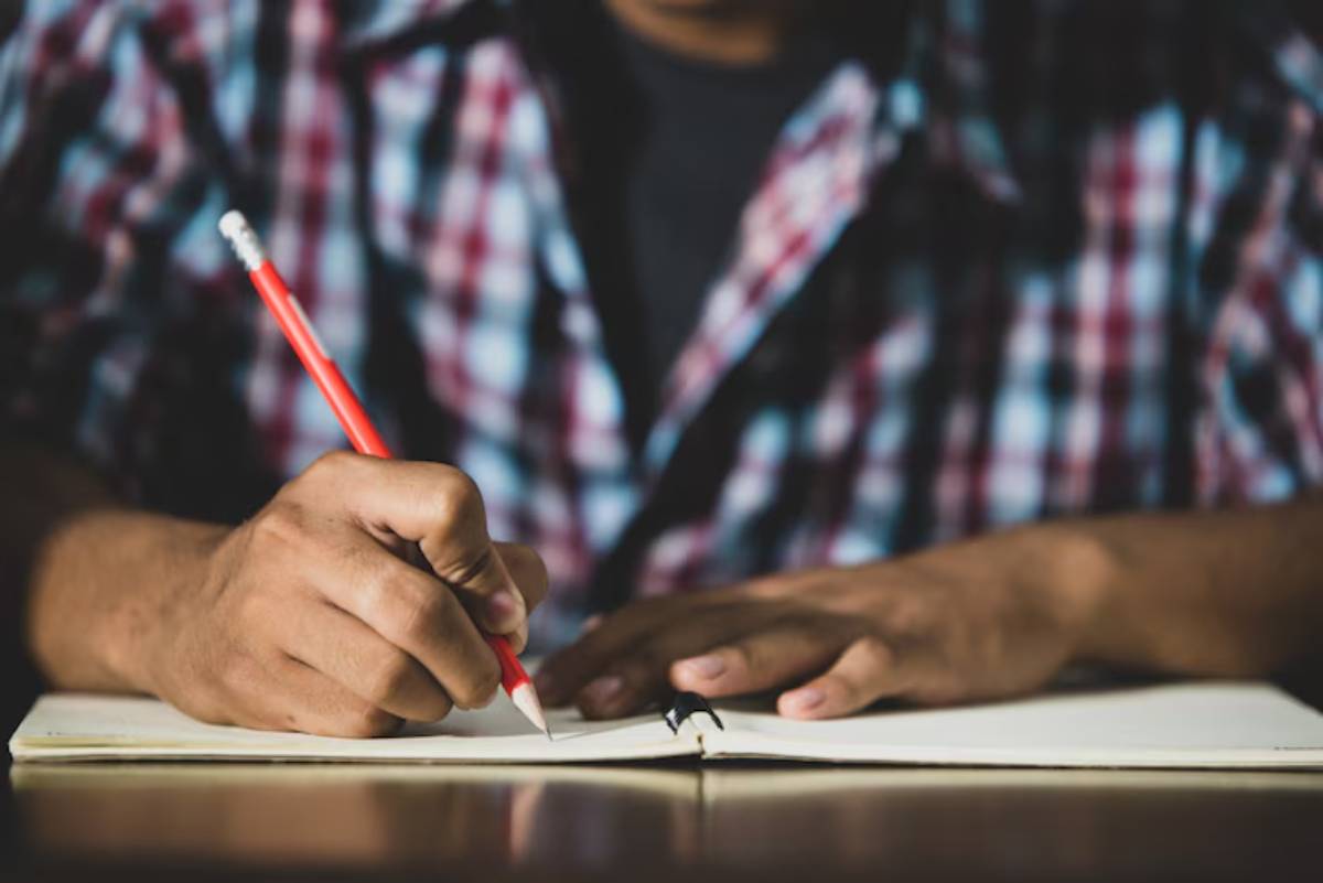 A person in a checkered shirt is writing in a notebook with a red pencil, focusing intently on their task.