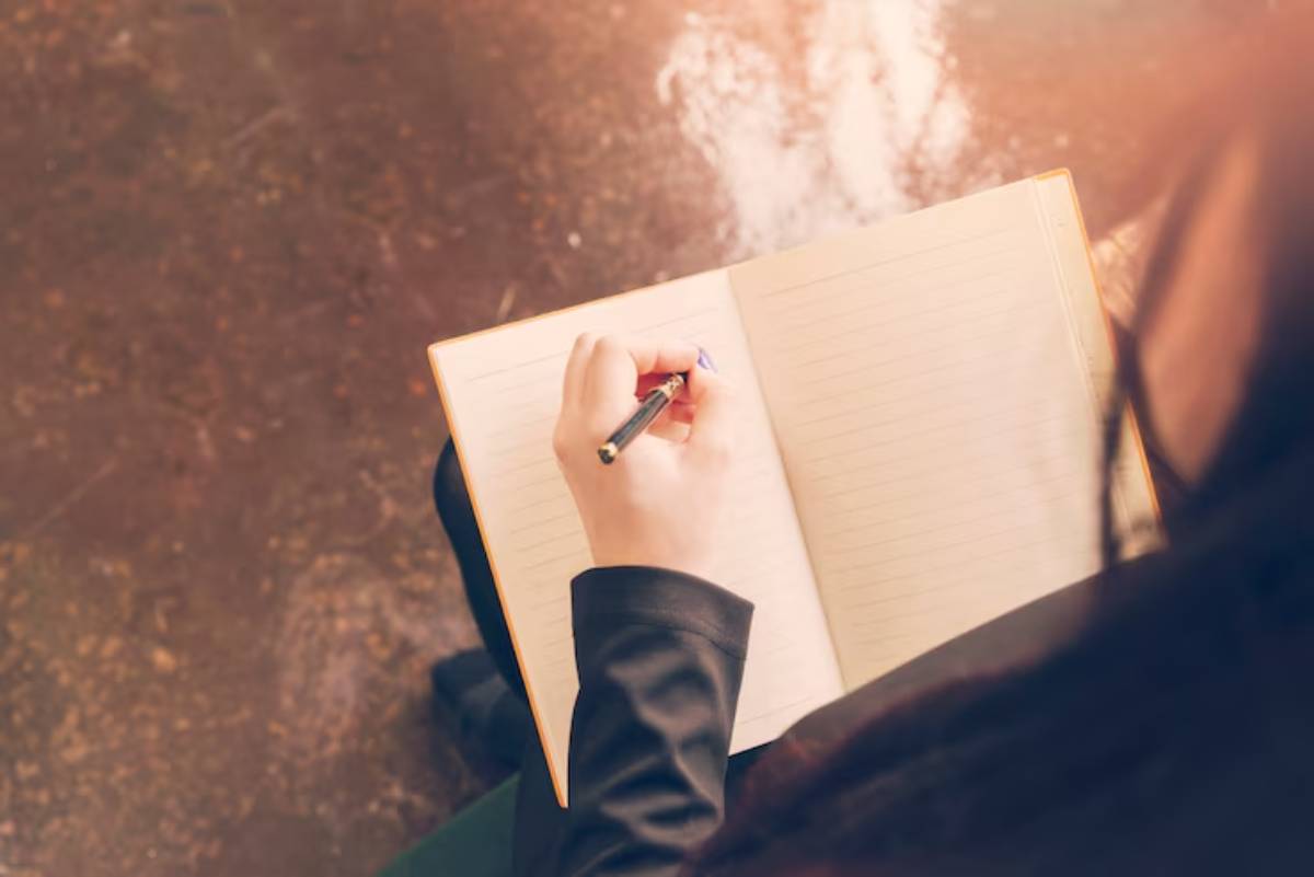 A person with long hair writes in a blank notebook with a blue pen, seated against a textured floor background.